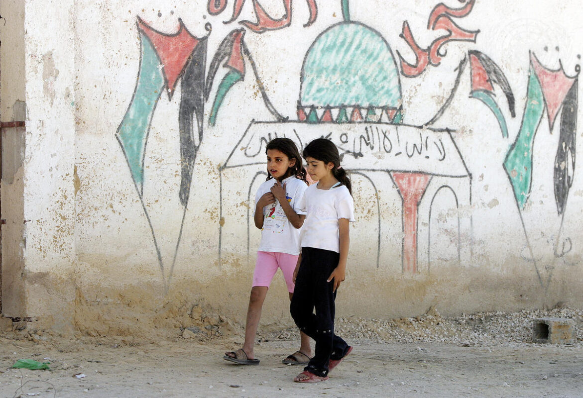 Two girls walk past a graffiti-strewn wall in the United Nations Relief and Works Agency for Palestine Refugees in the Near East (UNRWA)'s Acqba Jaber camp for Palestinian refugees, in the West Bank, located just outside of Jericho [UN Photo/Stephenie Hollyman]